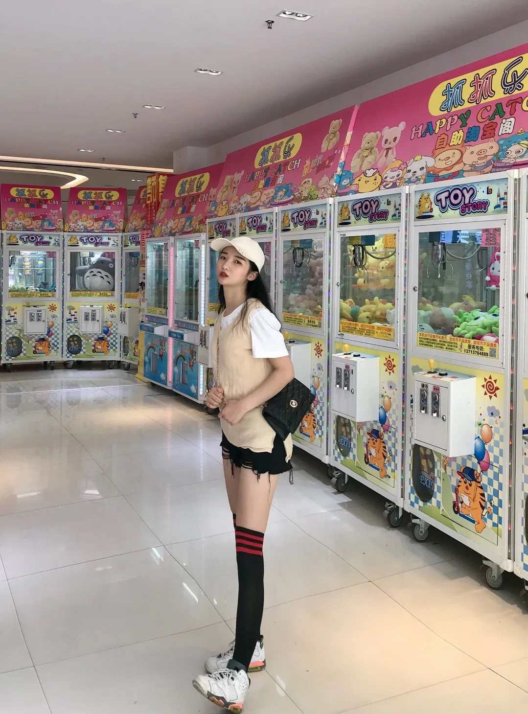 室内游艺厅彩绘抓娃娃机排区前身穿米色无袖针织背心、黑色短裤与中筒袜的站立半身像