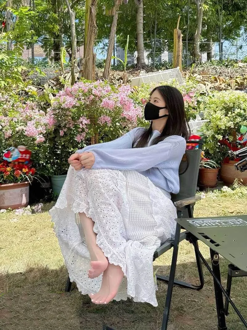 户外花园草坪旁,模特身着浅紫色开衫与白色镂空长裙赤足坐在露营折叠椅上