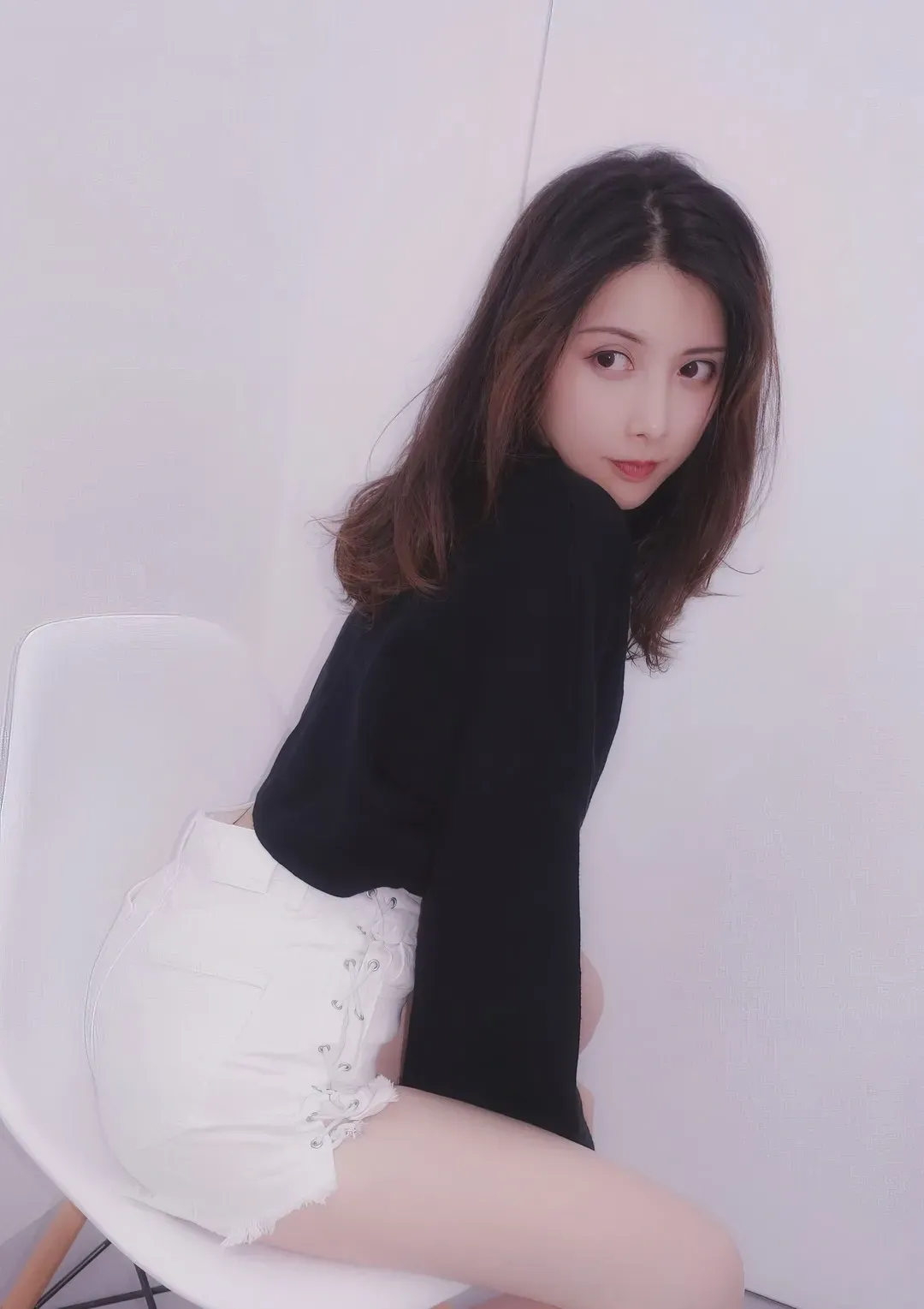 纯白室内背景下身穿黑色高领长袖上衣与白色系绳短裤侧坐白椅的女性中景